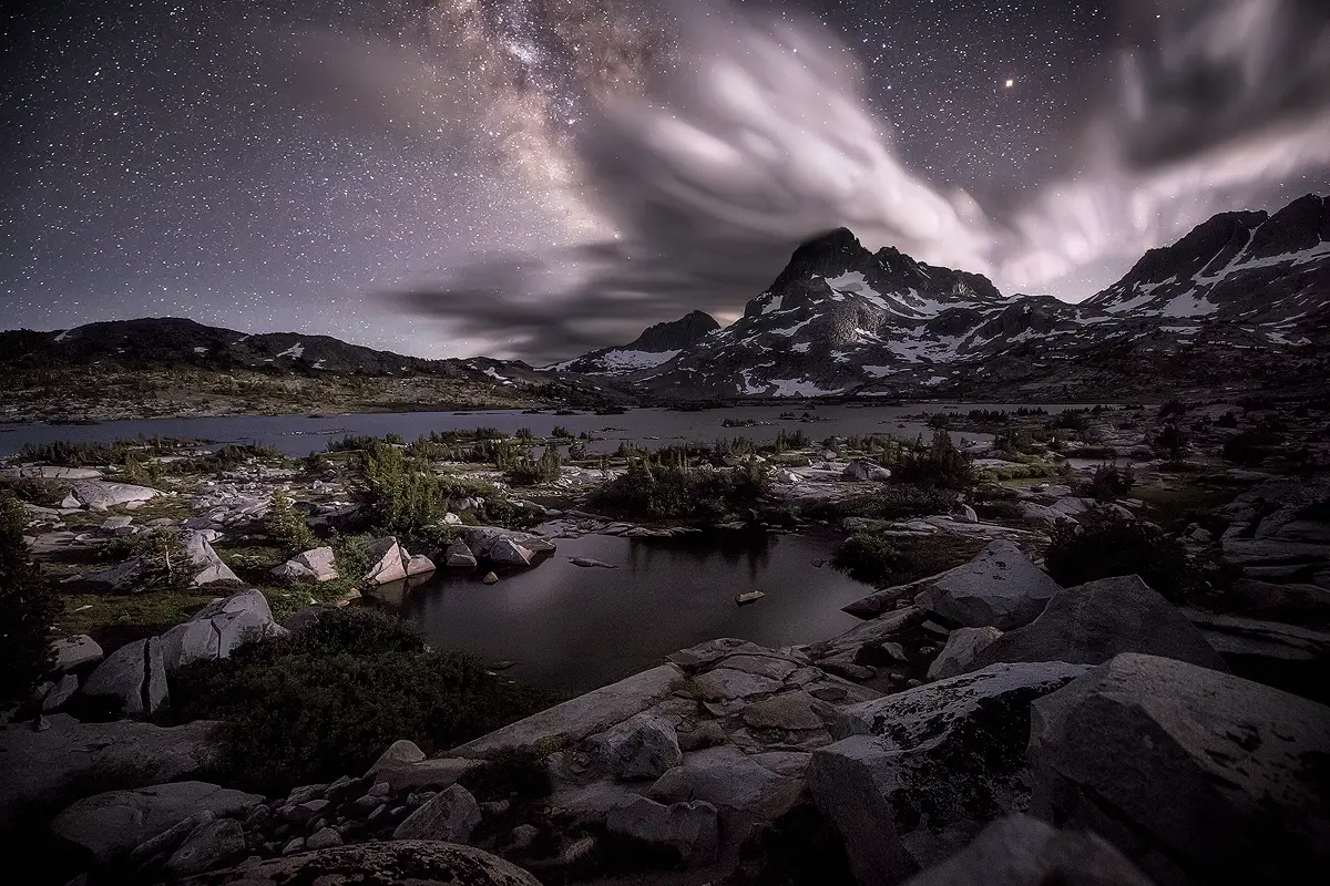 Milky Way core glows above a moonlit alpine lake and jagged peaks with streaking clouds overhead.