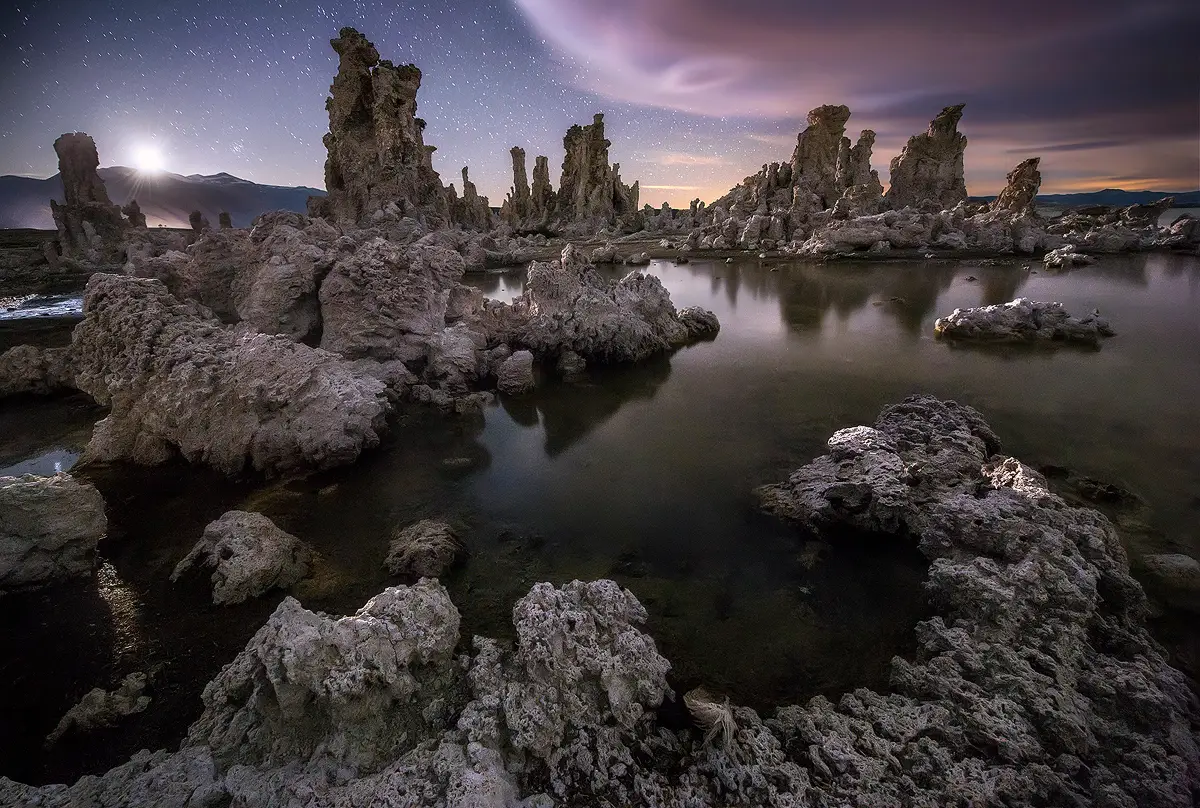 Moonlit tufa limestone towers rise from the still water of Mono Lake beneath a faint starry sky during moonset. 
