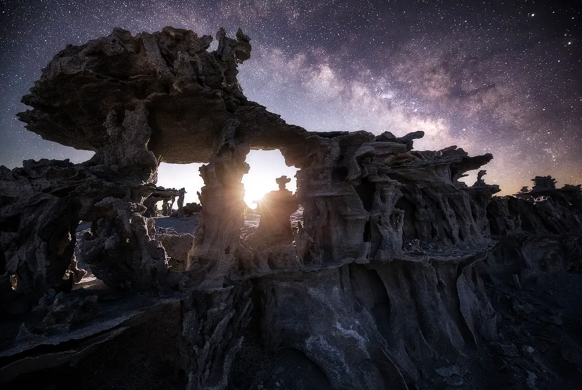 Milky Way galactic core during moonrise glowing behind a weathered sandstone arch formation under a star-filled sky.