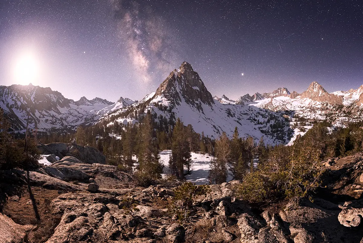 Rising moon illuminates a snow-covered alpine peak and granite basin under the Milky Way in the high Sierra.