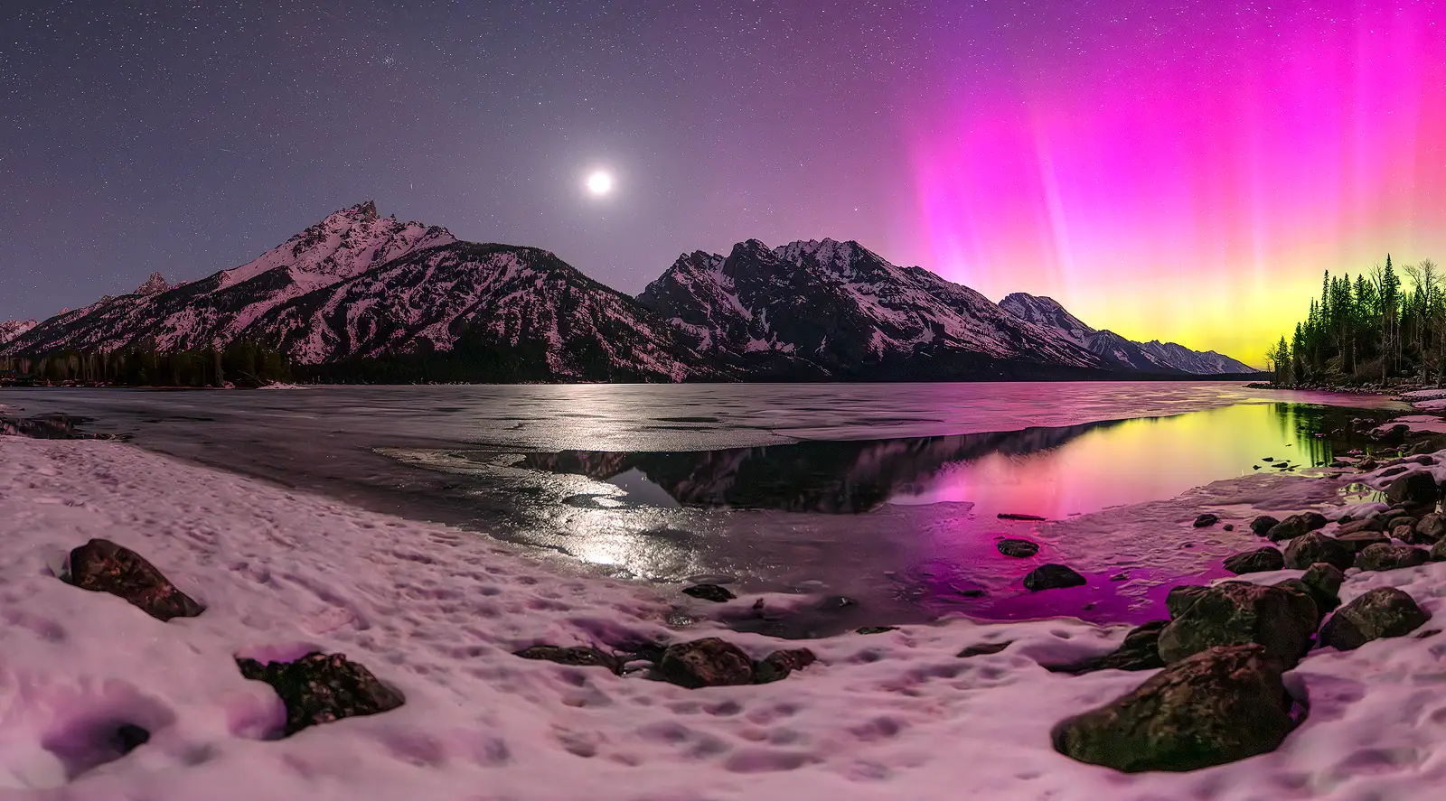 Pink and green aurora borealis dances beside the Grand Teton Range with a rising moon reflecting on a partially frozen lake