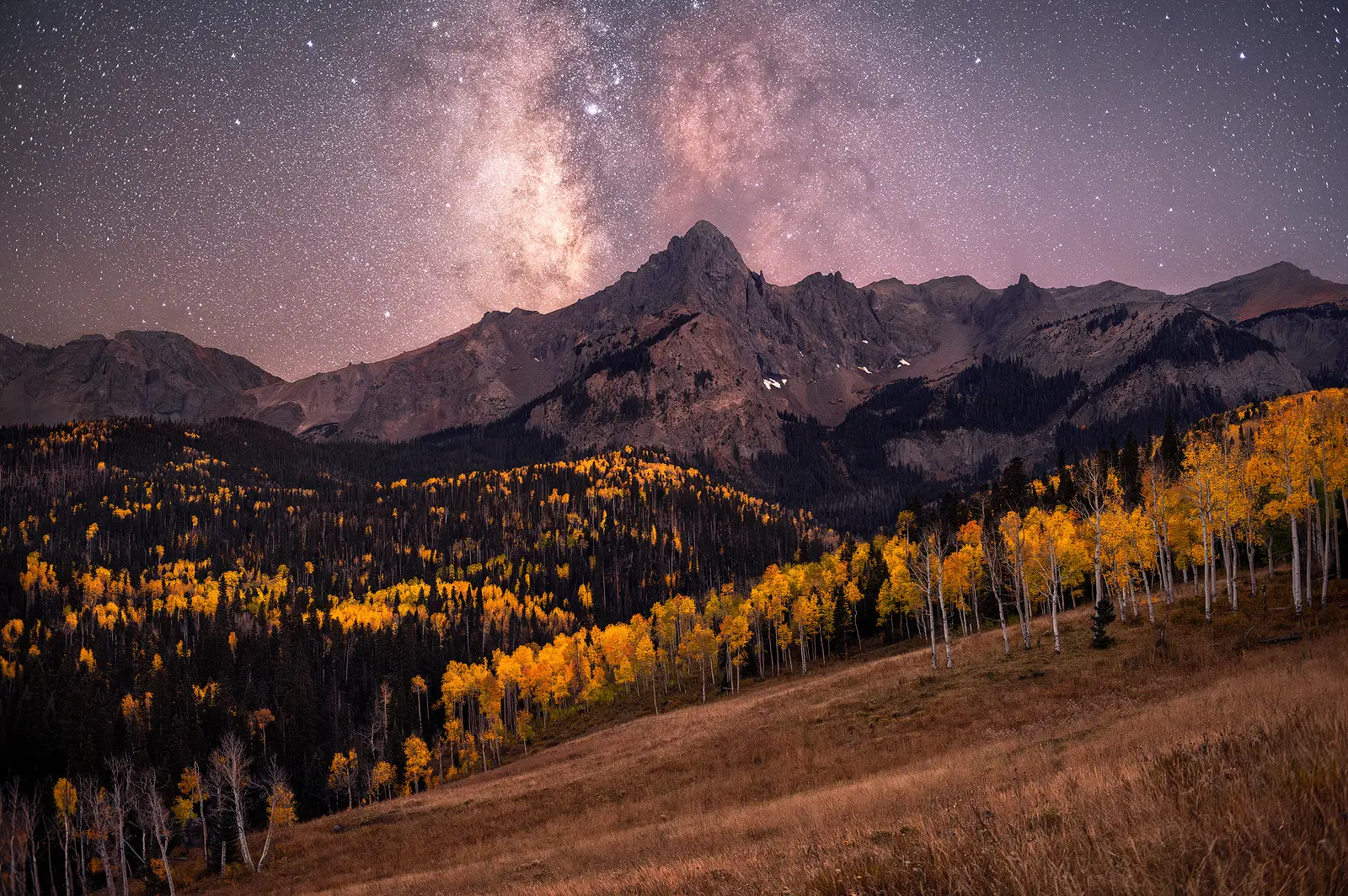 Milky Way core rising over a jagged Colorado peak with golden aspen groves illuminated in the foreground during autumn