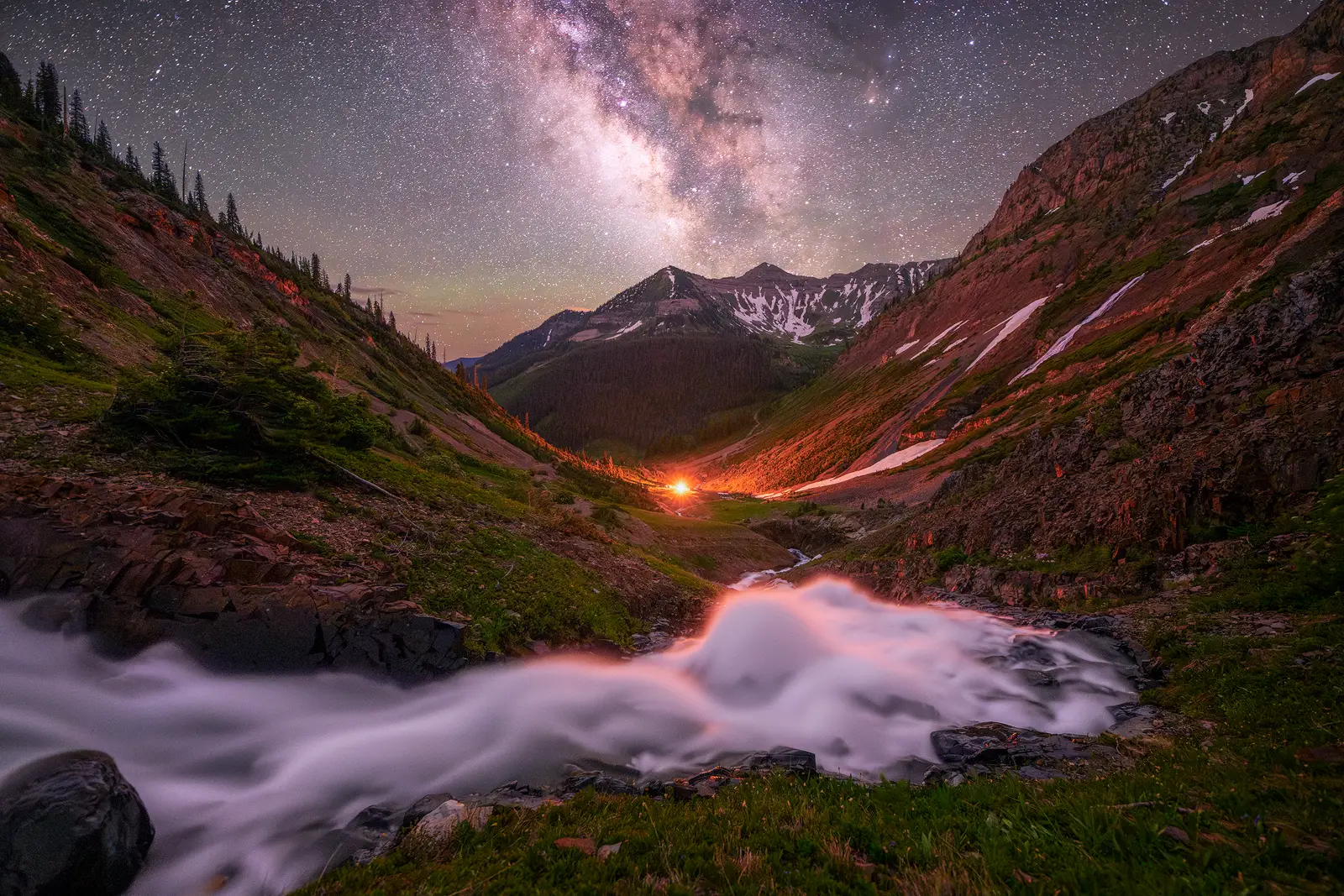 The Milky Way core rises above a Colorado Rockies valley with a distant campfire glowing in the mountains and a rushing creek in the foreground