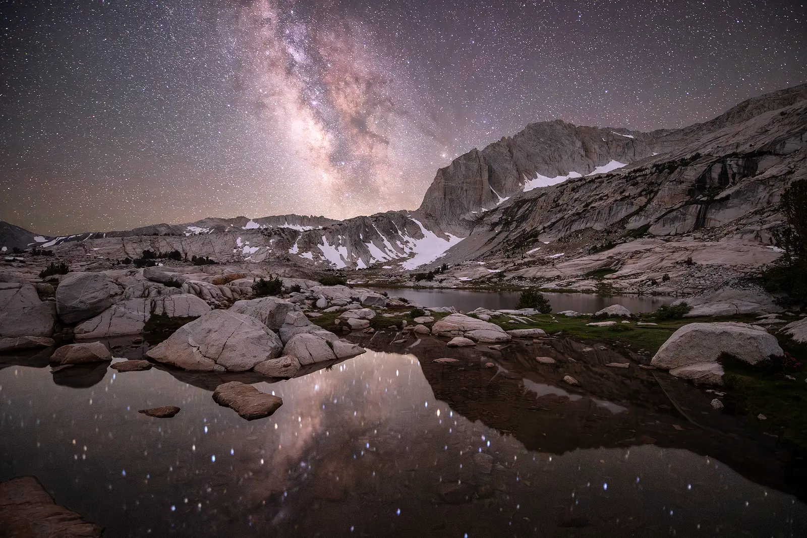 The Milky Way core reflects in a calm alpine lake surrounded by granite boulders and snow patches in the High Sierra Nevada