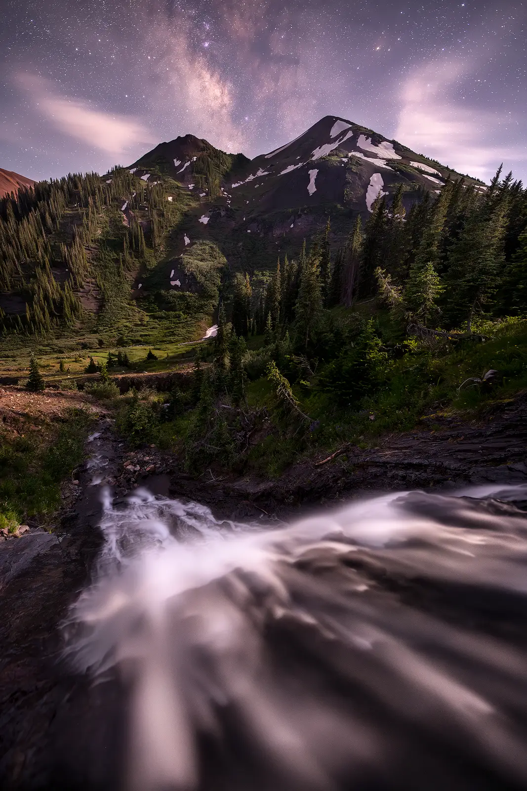 Moonlight illuminates a rushing waterfall and evergreen forest below a Colorado Rockies peak with the Milky Way visible through drifting clouds