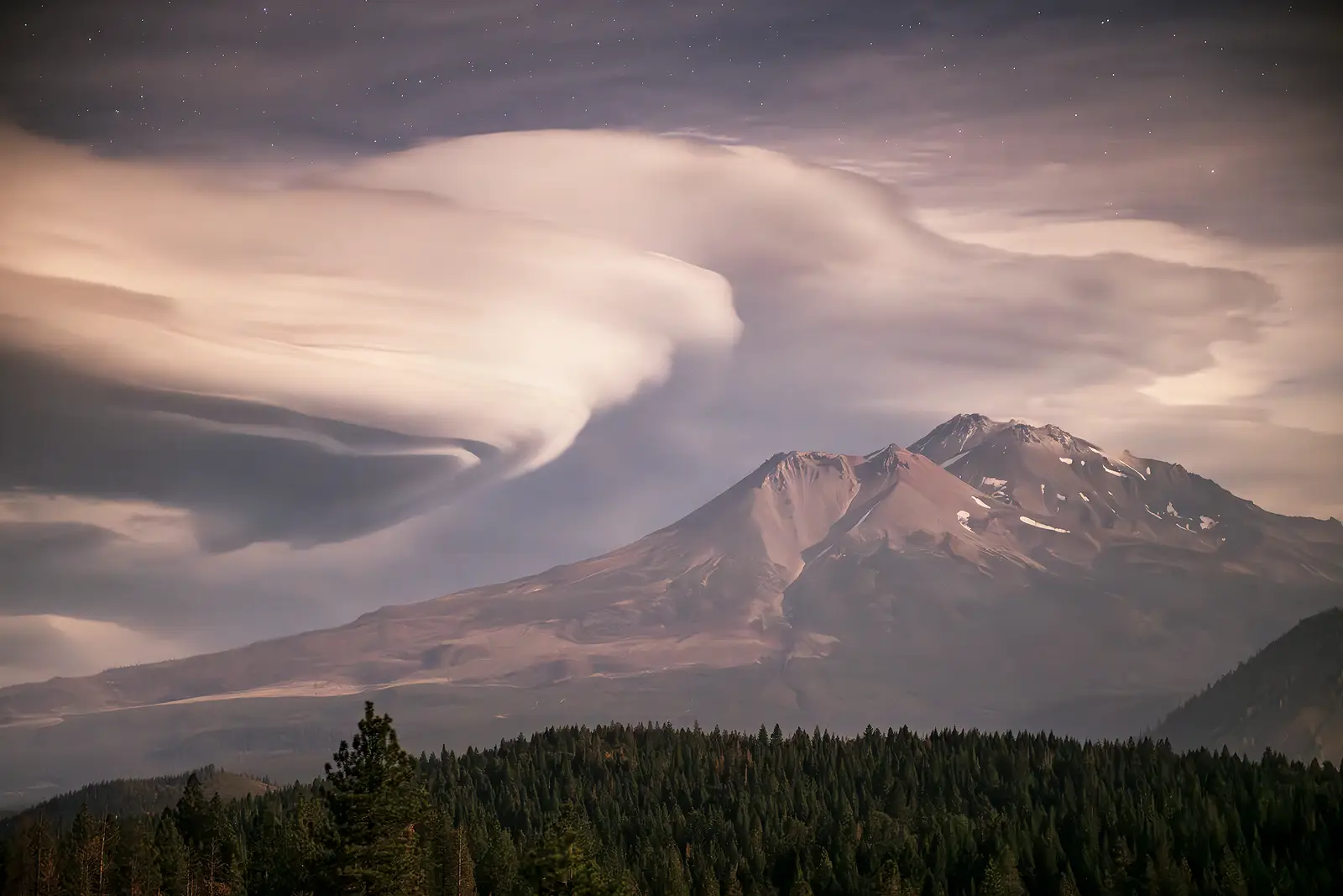 Moonlight illuminates swirling lenticular clouds forming above Mount Shasta with stars visible through breaks in the cloud cover