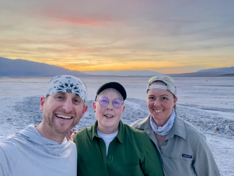 Brad Goldpaint and Evie Wilder pose with a workshop participant under a soft, colorful sky during sunset.