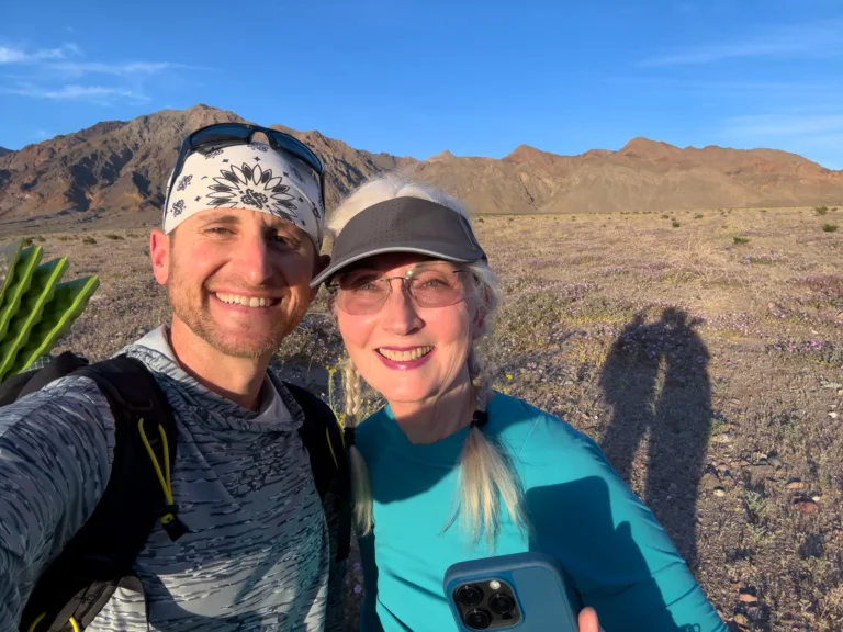 Brad Goldpaint and DJ taking a selfie among purple desert wildflowers with the mountain peaks of Death Valley behind them.