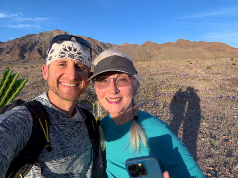 Brad Goldpaint and DJ taking a selfie among purple desert wildflowers with the mountain peaks of Death Valley behind them.
