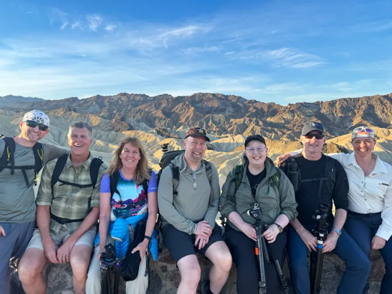 Brad Goldpaint and Evie Wilder pose with their night photography workshop participants seated together in front of the Zabriskie Point badlands under a blue sky.