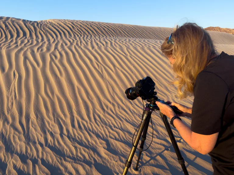 A night photography workshop participant adjusting a tripod-mounted camera to capture rippled dune patterns in warm evening light.