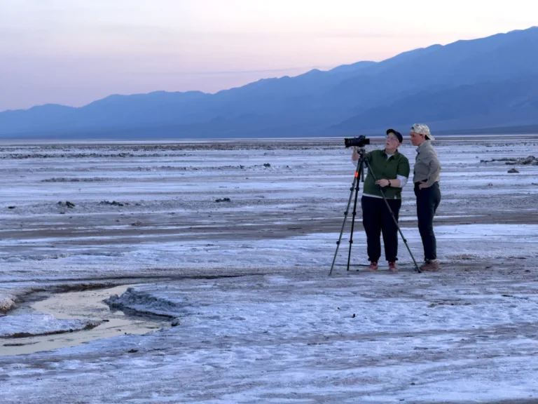 Evie Wilder helps a workshop participant set up their camera and discusses their composition on the salt flats under a soft pastel dusk sky.