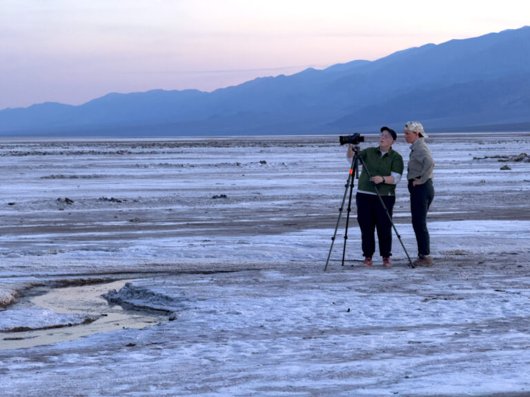 Evie Wilder helps a workshop participant set up their camera and discusses their composition on the salt flats under a soft pastel dusk sky.