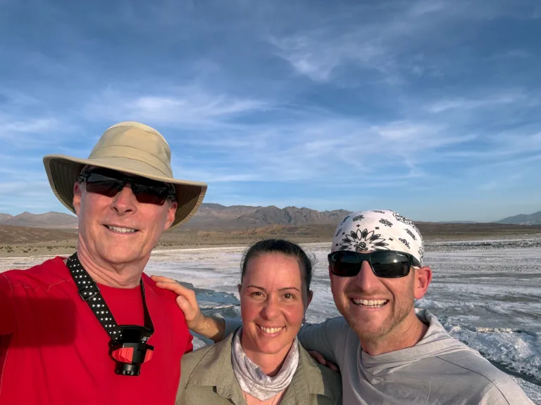 Brad Goldpaint and Evie Wilder pose with a night photography workshop participant on the Death Valley salt flats with mountains and a blue sky behind them.