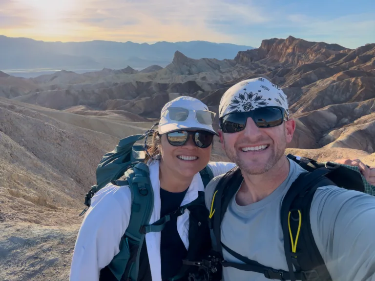 Brad Goldpaint and Kim pose for a selfie at Zabriskie Point, badlands and Manly Beacon behind them at sunset.